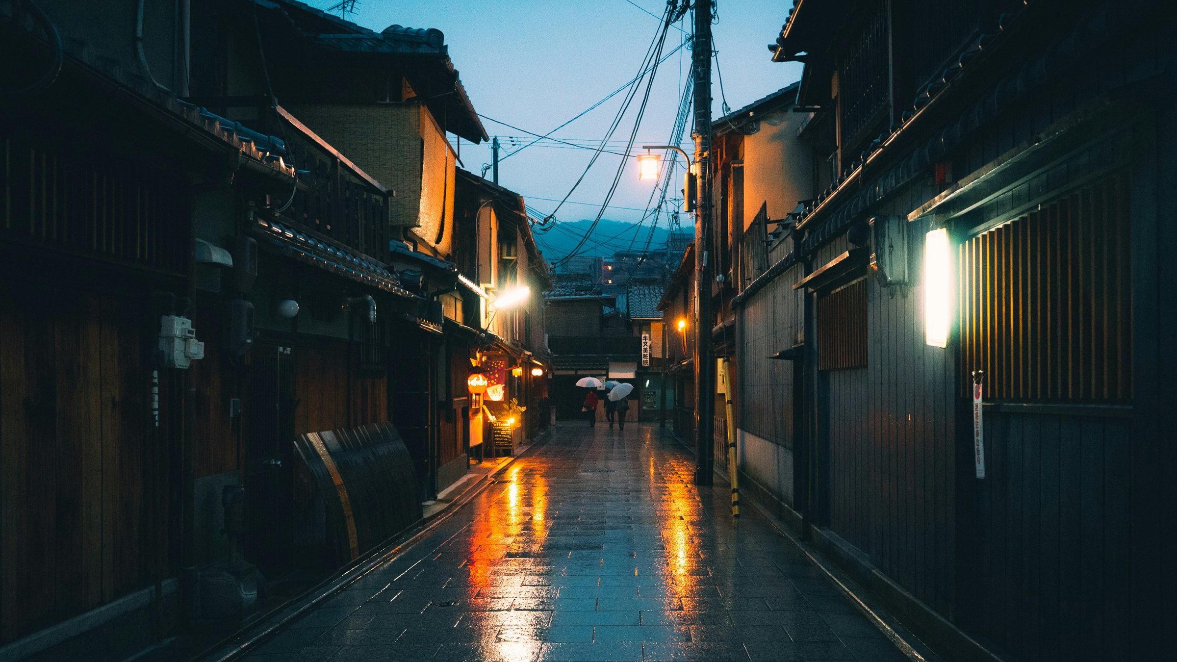 ซอยแคบปูหินเปียกฝนตอนพลบค่ำ มีบ้านไม้เรียงสองฝั่งและไฟถนนสีส้ม ไม่มีรถในภาพ